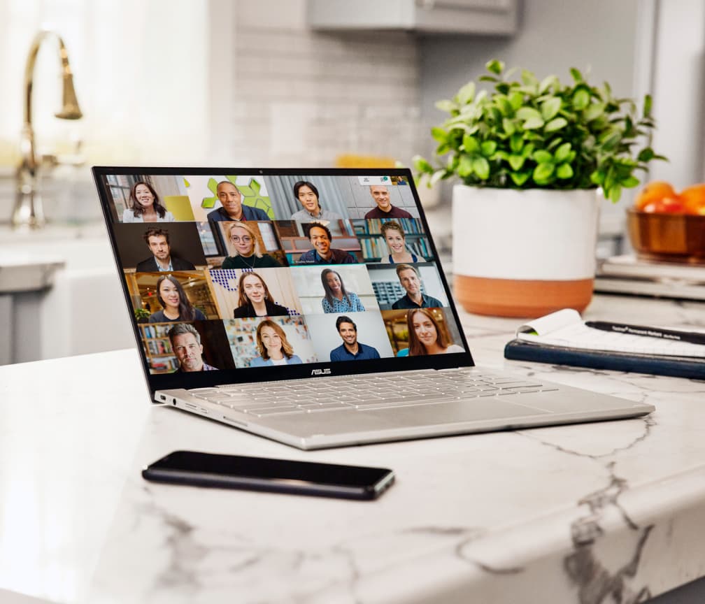 A Chromebook sits open on a white marble countertop with a cellphone on the left and a plant in the background.